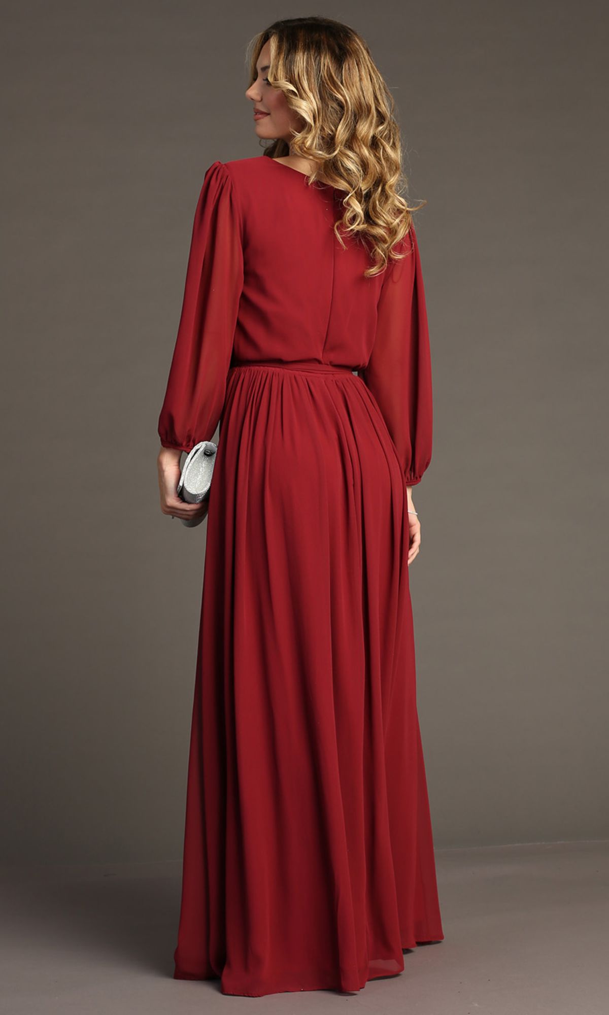 A woman stands with her back to the camera, wearing the Long-Sleeve Long Formal Dress: Chicas J012B in red and holding a silver clutch.